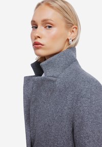 Grå uldfrakke med høj krave, der har en blød, tekstureret overflade. Modellens ansigt er delvist synligt, hvilket fremviser klar hud og let makeup.