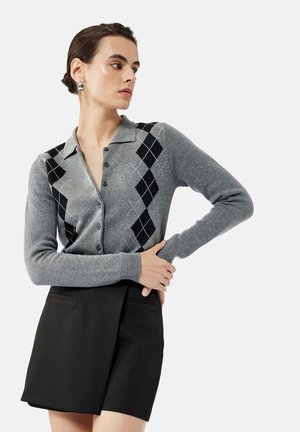 Harmaa neulecardigan, jossa on musta timantinmuotoinen kuvio, napit edessä ja pehmeä rakenne. Yhdistetty mustaan lyhyeen hameeseen, jossa on etutaskut.