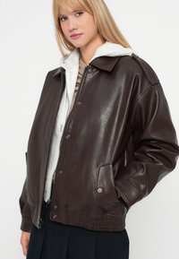 Veste en simili cuir marron avec une coupe décontractée, boutons-pression, deux poches avant et un sweat à capuche blanc en dessous, présentant une finition texturée.