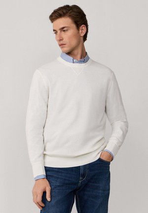 Un pull blanc avec un design en V, fait d'un matériau doux. Associé à une chemise collée rayée bleue qui dépasse des poignets.