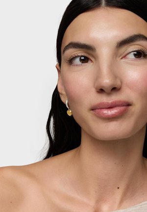 Donna con capelli scuri che indossa orecchini a cerchio d'argento con piccoli ciondoli a forma di cuore dorato, che guarda leggermente verso destra, spalle scoperte visibili.