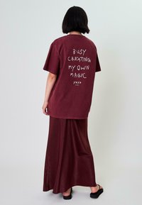 Bordové oversized tričko s bílým nápisem "USILOVNĚ TVOŘÍM SVOU VLASTNÍ MAGII" na zadní straně. V páru s odpovídající bordovou maxi sukni. Hladká textura.
