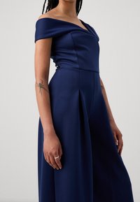 Mono azul marino con escote off-the-shoulder, cuerpo ajustado, pliegues relajados en la cintura y tejido suave y texturizado.