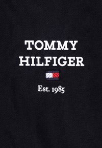 Tela negra con texto bordado en blanco: "TOMMY HILFIGER" y "Est. 1985", con un pequeño logo en rojo, blanco y azul debajo.