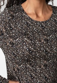 Brun och svart mönstra tyg med intrikata blommiga och abstrakta mönster. Rundad halsringning och mjuk textur. Närbild.