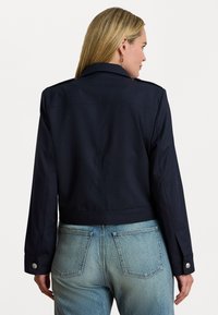 Giacca cropped blu navy con maniche lunghe, colletto a rever e dettagli in bottone argentato, abbinata a jeans in denim azzurro. Tessuto liscio e testurizzato.