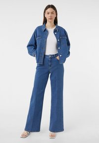 Blauwe spijkerjasje met twee voorzakken, in combinatie met hoge blauwe spijkerjeans met wijde pijpen. Wit geribbeld topje eronder en witte sandaaltjes met bandjes.