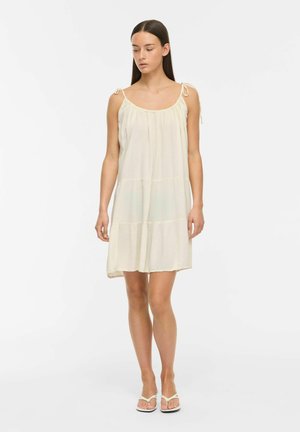 Jeune femme aux longs cheveux foncés portant une robe crème ample, sans manches, avec des bretelles nouées et des sandales blanches, debout devant un fond uni.