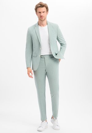 Homme portant un costume vert clair avec un t-shirt blanc et des baskets blanches, debout sur fond blanc.