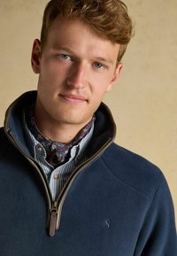 Mladý muž so svetlo hnedými vlasmi, oblečený v námornícky polar fleece, zapnutý do polovice cez pruhovanú košeľu a s kvetinovou šatkou okolo krku, na béžovom pozadí.