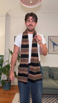 Kötött sál barna, fekete és olíva csíkokkal, puha tapintással. Az egyén mikrofont tart, fehér pólót és farmert visel.