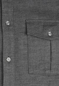 Camicia grigia con bottoni in tessuto texturizzato, dotata di due bottoni in argento e una tasca sul petto. Il design presenta un sottile motivo diagonale.
