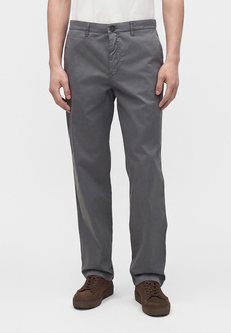 Homme portant un pantalon gris droit et des chaussures marron à lacets, debout devant un fond uni.