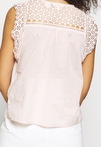 Haut rose clair avec un délicat panneau en dentelle sur les épaules, des bords festonnés et un tissu fluide et doux.