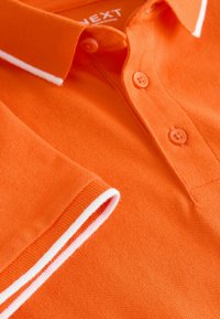 Polo orange en tissu texturé, avec un col rayé blanc et des boutons assortis.