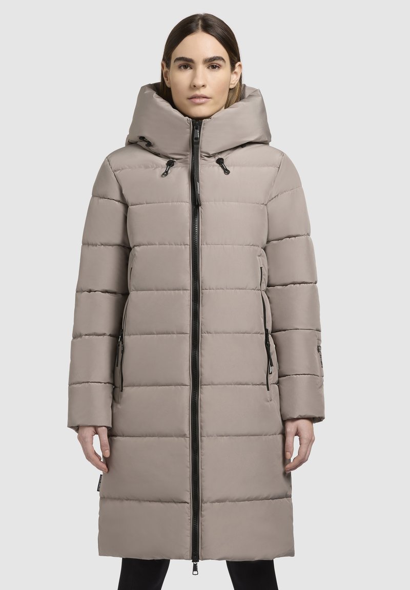 Beige dunjakke med hætte, med horisontalt quiltet design, lynlåslukning foran og sidelommer med sorte lynlåse.
