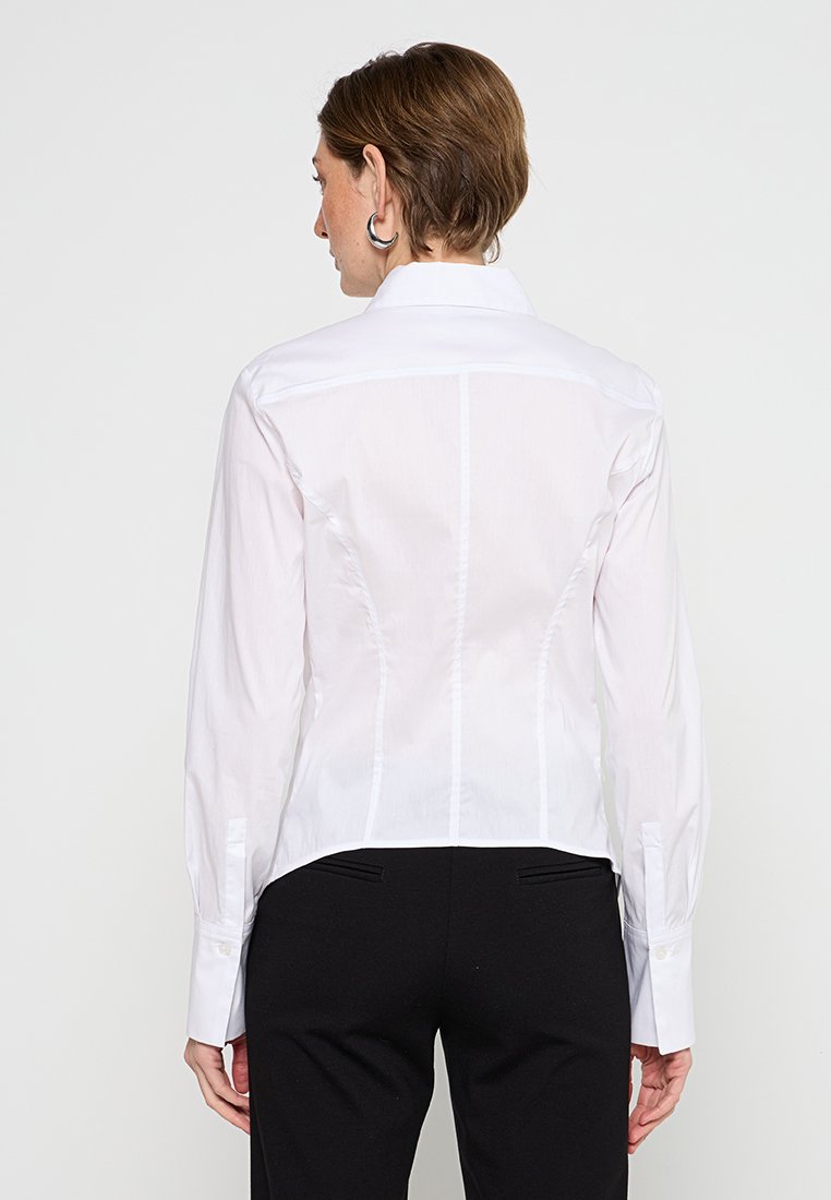 Person mit kurzen braunen Haaren, der ein enganliegendes weißes Langarmshirt und schwarze Hosen trägt, steht mit dem Rücken zu einem schlichten Hintergrund.