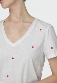 T-shirt blanc à col en V fabriqué dans un tissu doux avec des cœurs rouges brodés éparpillés. Manches courtes, design minimaliste, coupe décontractée.