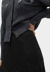 Grå ullkofta med ribbade muddar och dekorativa ädelstenar, kombinerad med en svart spetskjol med blommönster och volangkant.