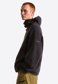 Σκούρο γκρι φλις φούτερ με ψηλό γιακά, μακριά μανίκια και τσέπη με φερμουάρ στον άνω βραχίονα. Το ύφασμα φαίνεται μαλακό και με υφή.