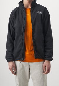 Schwarze Fleecejacke mit Reißverschluss, hohem Kragen und Logo auf der linken Brust. Kombiniert mit einem orangefarbenen Shirt darunter.