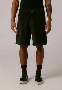 Man wearing black corduroy shorts, black socks, and black slip-on sneakers, standing with arms relaxed by sides.
