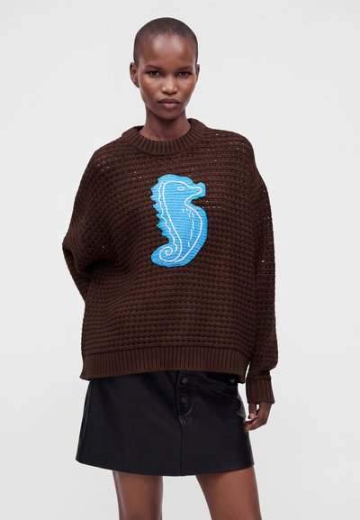 Femme portant un pull en tricot marron avec un grand motif hippocampe bleu et une jupe en cuir noire boutonnée sur un fond uni.