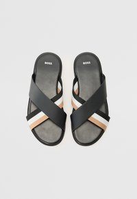 Svarta sandaletter med korsade remmar. Remmarna har beige, vita och svarta accenter. Platt sula med en mjuk textur och logotyp.