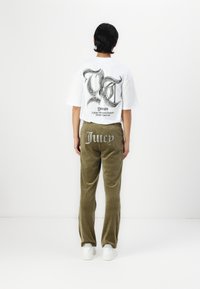 Weißes T-Shirt mit großem schwarzem Grafikdesign auf dem Rücken, kombiniert mit olivgrünen Cordhosen, die "Juicy" in Strasssteinen tragen. Weiße Sneaker.