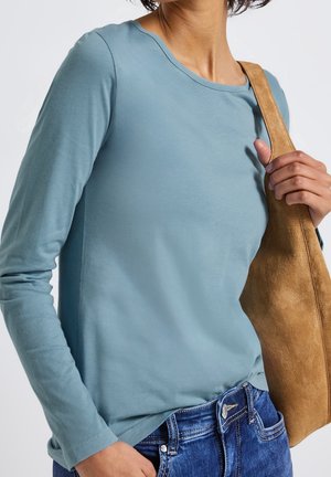 Femme portant une chemise à manches longues bleu clair et un jean bleu, tenant la sangle d'un sac à épaule en daim beige de la main droite.