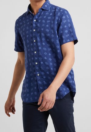 Mężczyzna w granatowej koszuli z krótkim rękawem zapinanej na guziki, z jasnoniebieskimi wzorami sportowymi, i ciemnych spodniach na neutralnym tle.