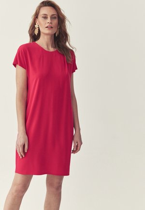 Femeie cu păr lung purtând cercei cu frunze de aur și o rochie simplă roșie cu mâneci scurte, pe un fundal deschis.