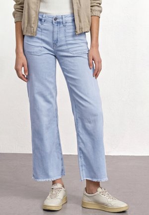 Personne portant un jean large bleu clair avec des ourlets effilochés, une veste beige et des baskets écru, debout contre un mur blanc.