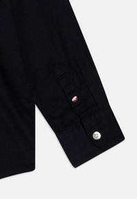 Camicia a maniche lunghe di colore blu scuro con tessuto testurizzato e polsini con bottoni. Presenta un piccolo accento rosso e bianco sui bottoni dei polsini.