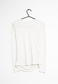Chemise blanche à manches longues en tissu extensible et lisse. Dotée d'un col rond et d'un ourlet arrondi à l'arrière. Suspendue à un crochet noir.