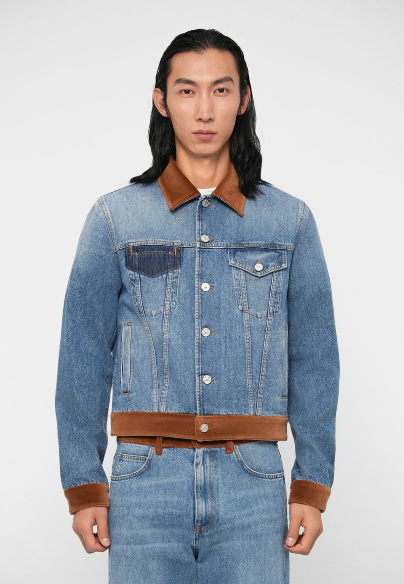 Chaqueta de mezclilla en azul claro con un cuello y puños de ante marrón. Presenta bolsillos frontales y botones de metal en la paleta y los puños.