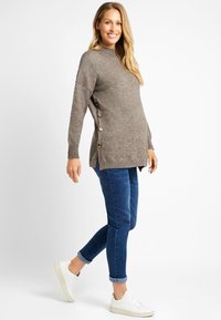 Pull en maille brun clair avec des bords côtelés, des boutons sur les côtés et un col haut, associé à un jean bleu foncé et des baskets blanches.