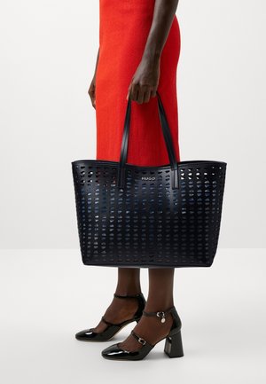 Persoon in een rode jurk die een grote zwarte geperforeerde draagtas met het logo "HUGO" vasthoudt, draagt zwarte enkellaarsjes met een bandje van lakleer.