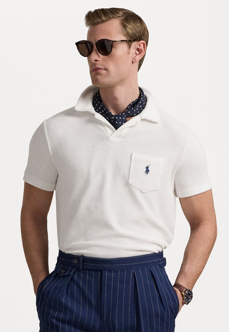 Polo shirt blanc avec une poche et un logo bleu, associé à un pantalon rayé navy. Comprend des lunettes de soleil noires et un foulard à pois.
