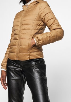 Personne portant une veste matelassée beige avec fermeture éclair et un pantalon en cuir noir, une main dans la poche de la veste, debout devant un fond uni.