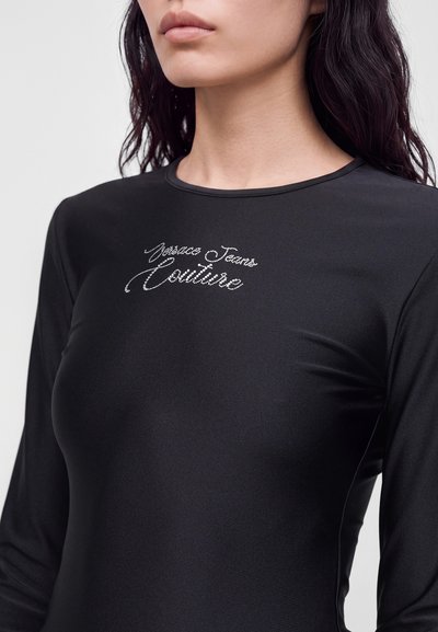 Femme portant un haut noir ajusté à manches longues avec des lettres scintillantes "Versace Jeans Couture" sur la poitrine.