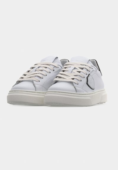 Baskets en cuir blanc avec lacets blancs et semelles épaisses en caoutchouc, avec un détail en patch noir sur le côté sur un fond uni.