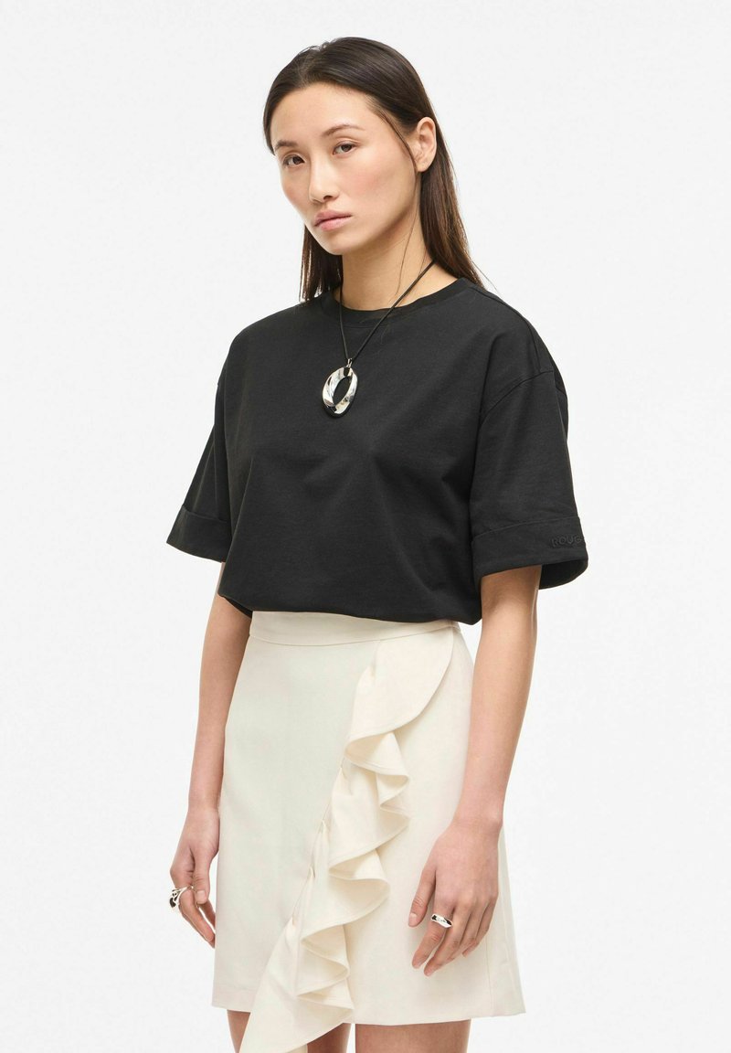 Jeune femme portant un T-shirt noir oversize, une jupe blanche avec un volant latéral, un collier argenté et des bagues, debout devant un fond uni.