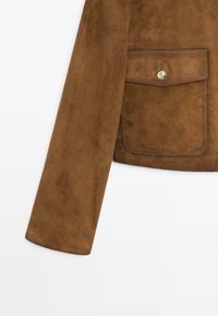 Manica di giacca in suede marrone e tasca a pattina con bottone argentato su sfondo bianco, che mostra dettagli di cucitura fine.