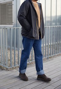 Giacca nera in nylon con dettagli gialli, maglione beige in maglia, jeans in denim blu e stivali marroni con lacci. In piedi su una superficie di legno.