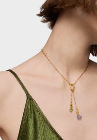 Złoty naszyjnik łańcuszkowy z podwójnym splotem, ozdobiony trójkątnym zielonym kamieniem i okrągłym fioletowym kamieniem, na szyi modelki.