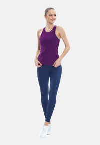 Giovane donna in piedi, indossa un top senza maniche viola, leggings blu scuro e sneakers bianche, guarda di lato sorridendo.