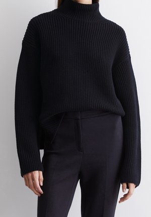 Frau trägt einen schwarzen, gerippten Rollkragenpullover mit langen Ärmeln und schwarze, enganliegende Hose, steht vor einem schlichten Hintergrund.