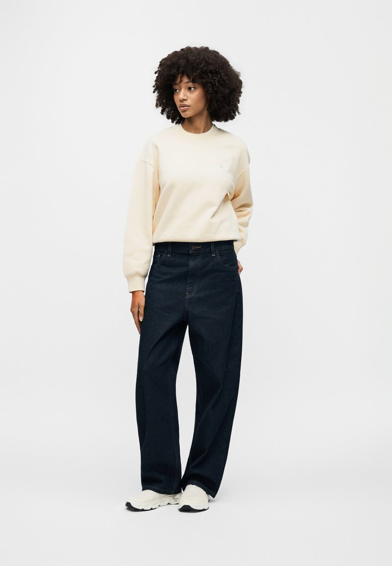 Cremefarbener Sweatshirt mit kleinem Logo, lockere dunkle Jeans und weiße Sneaker. Das Model steht mit neutraler Haltung vor einem weißen Hintergrund.