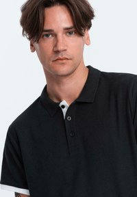 Černé polo tričko s bílým límcem. Obsahuje dvě knoflíky na léze a texturovanou látku. Model má vlasy střední délky.